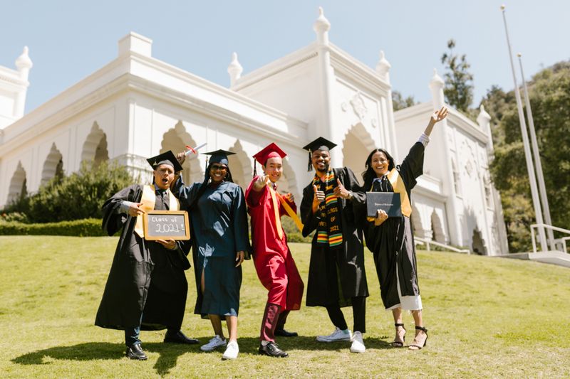 A group of diverse graduates posing joyfully wearing caps and gowns outside a historic building.